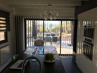 Vue depuis une cuisine avec îlot central sur un store californien installé sur une baie vitrée, lames orientables laissant passer la lumière et offrant une vue sur la terrasse et le jardin
